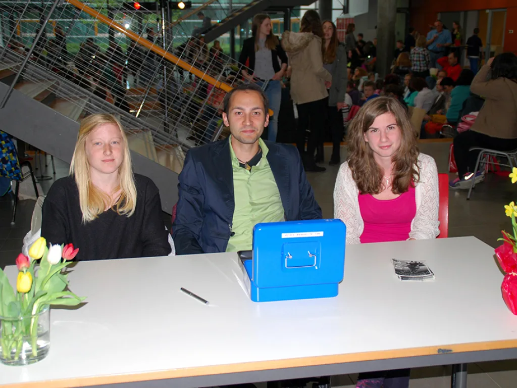 Drei Personen sitzen an einem Tisch mit Blumen und einer blauen Kasse.